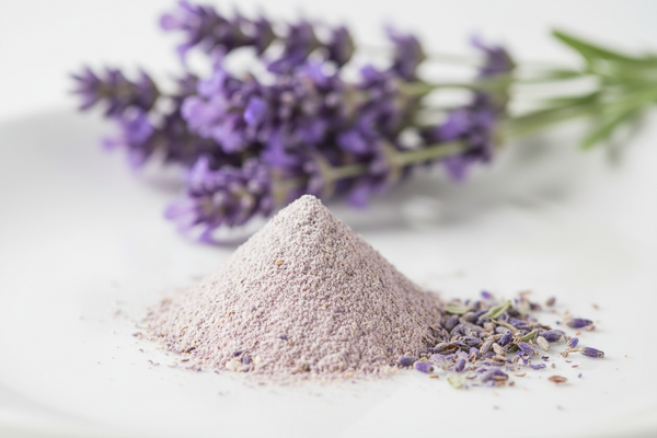 Create a premium ingredient photo of Lavender Extract.
Show a small amount of fine purple-tinted or off-white botanical powder on a clean surface, with a few lavender flowers softly blurred behind it.
Soft pastel lighting, calming, wellness-focused photography.